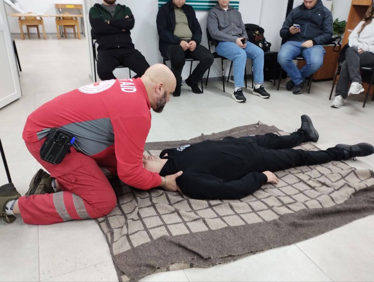 Інструктор у червоній формі з написом FIRST AID (Перша Допомога) схилився над манекеном або особою, що лежить на спині на ковдрі на підлозі, демонструючи техніку перевірки дихання або стану свідомості. Кілька людей спостерігають за демонстрацією, сидячи на стільцях позаду.