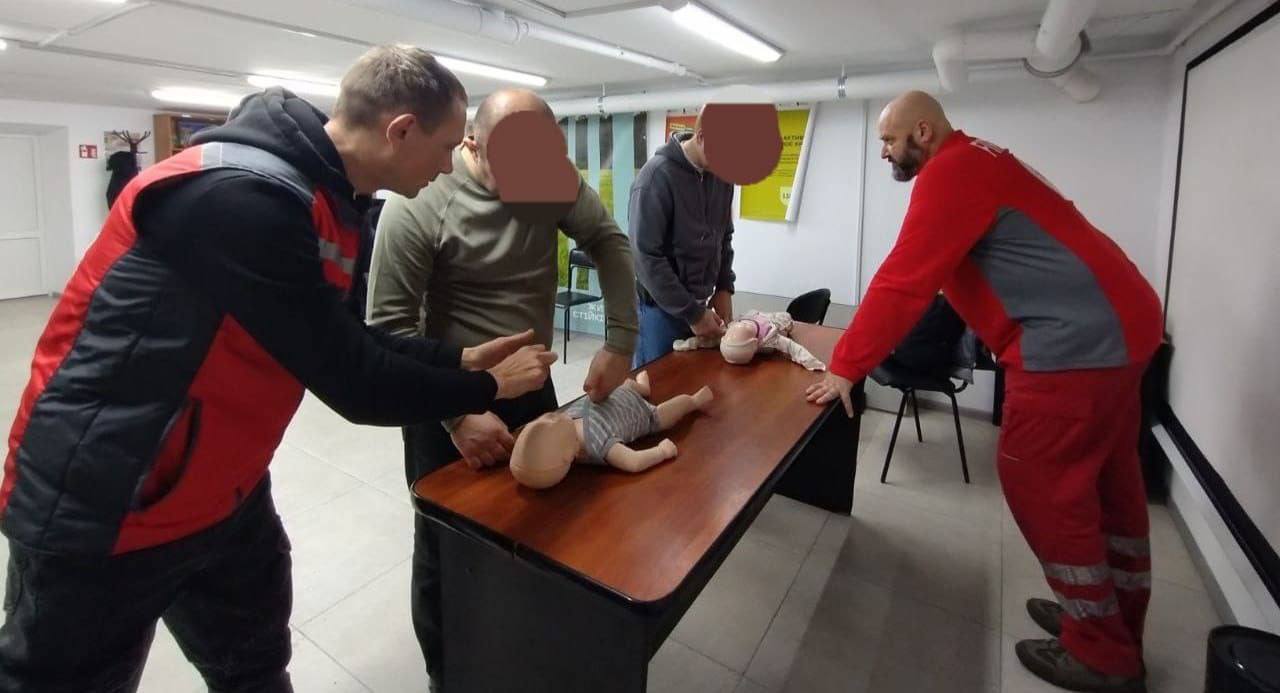 Четверо чоловіків у приміщенні беруть участь у тренінгу з першої допомоги. Двоє з них стоять ближче до манекена немовляти, що лежить на дерев'яному столі і відпрацьовують прийоми. Ще один чоловік у червоній формі інструктора стоїть збоку столу, спостерігаючи за процесом. На столі лежить другий манекен, схожий на дитину.