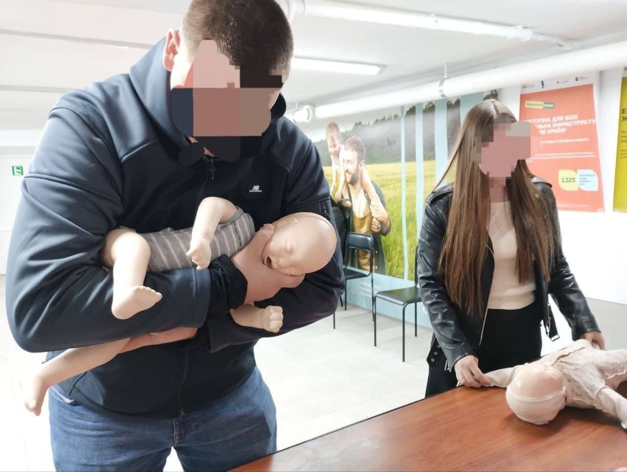 Чоловік у синій куртці та джинсах тримає на руках манекен немовляти, відпрацьовуючи техніку допомоги при задусі або інші реанімаційні дії. Жінка в чорній шкіряній куртці стоїть поруч, дивлячись на інший манекен дитини, що лежить на столі. Це сцена з практичного заняття з першої допомоги.