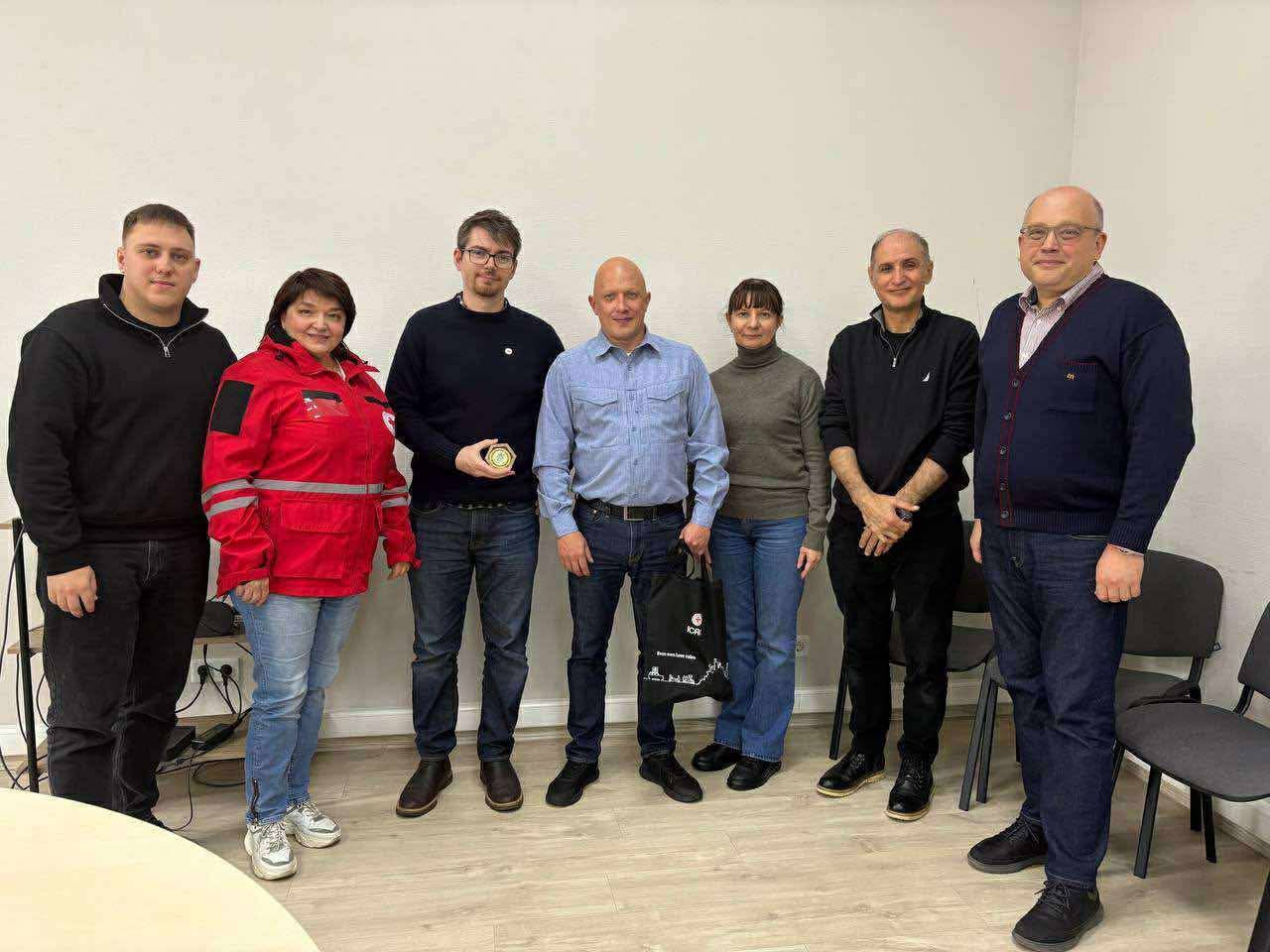 Групове фото семи людей у звичайному одязі, які стоять у світлій кімнаті. Серед них жінка в червоній куртці, схожій на форму Червоного Хреста, та чоловік, який тримає невеликий круглий предмет (медаль або значок).