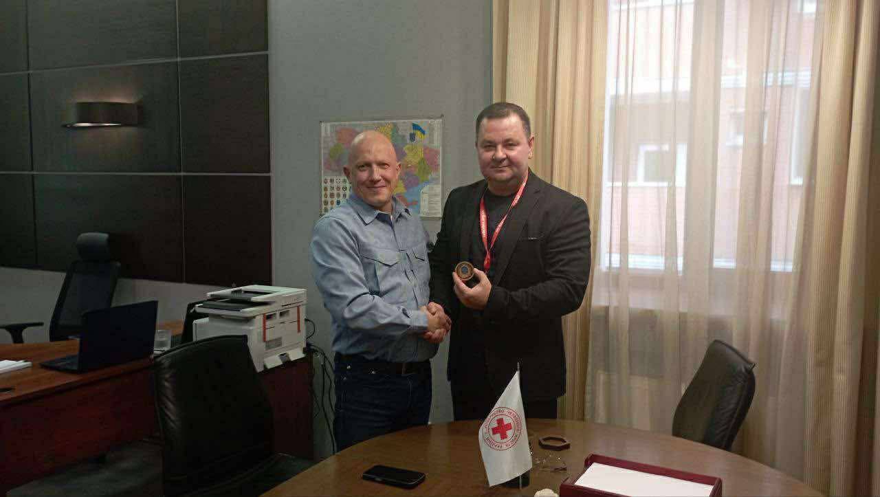 Двоє чоловіків потискають один одному руки в офісі. Чоловік у чорному піджаку та з медаллю на шиї тримає в руці круглий предмет. На столі перед ними стоїть маленький прапорець із символом Червоного Хреста.