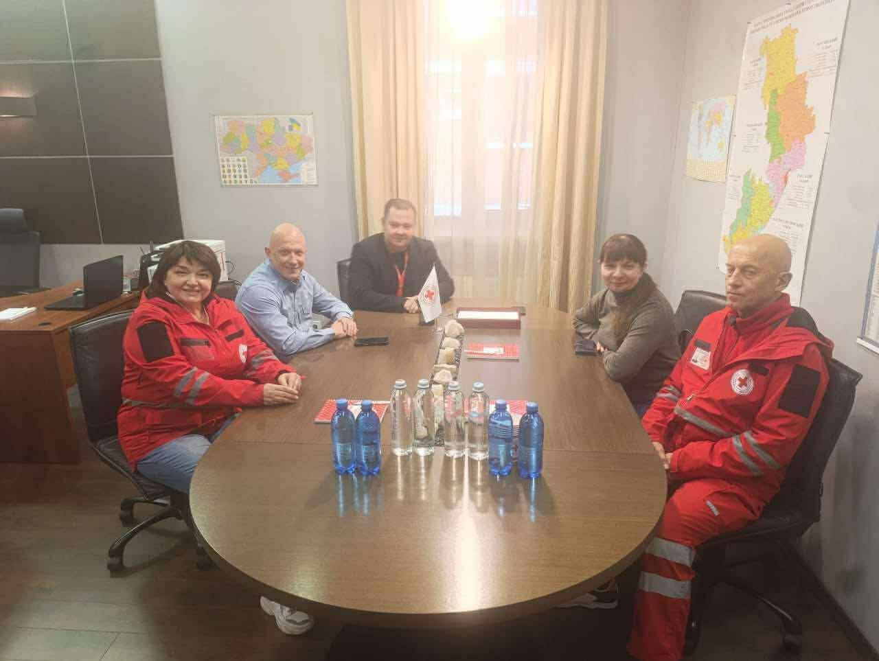 П'ятеро людей сидять за овальним столом у приміщенні для нарад. Дві особи (жінка ліворуч і чоловік праворуч) одягнені в червоні куртки, схожі на форму Червоного Хреста. На столі стоять пляшки з водою та маленький прапорець Червоного Хреста. На стінах видно карти.