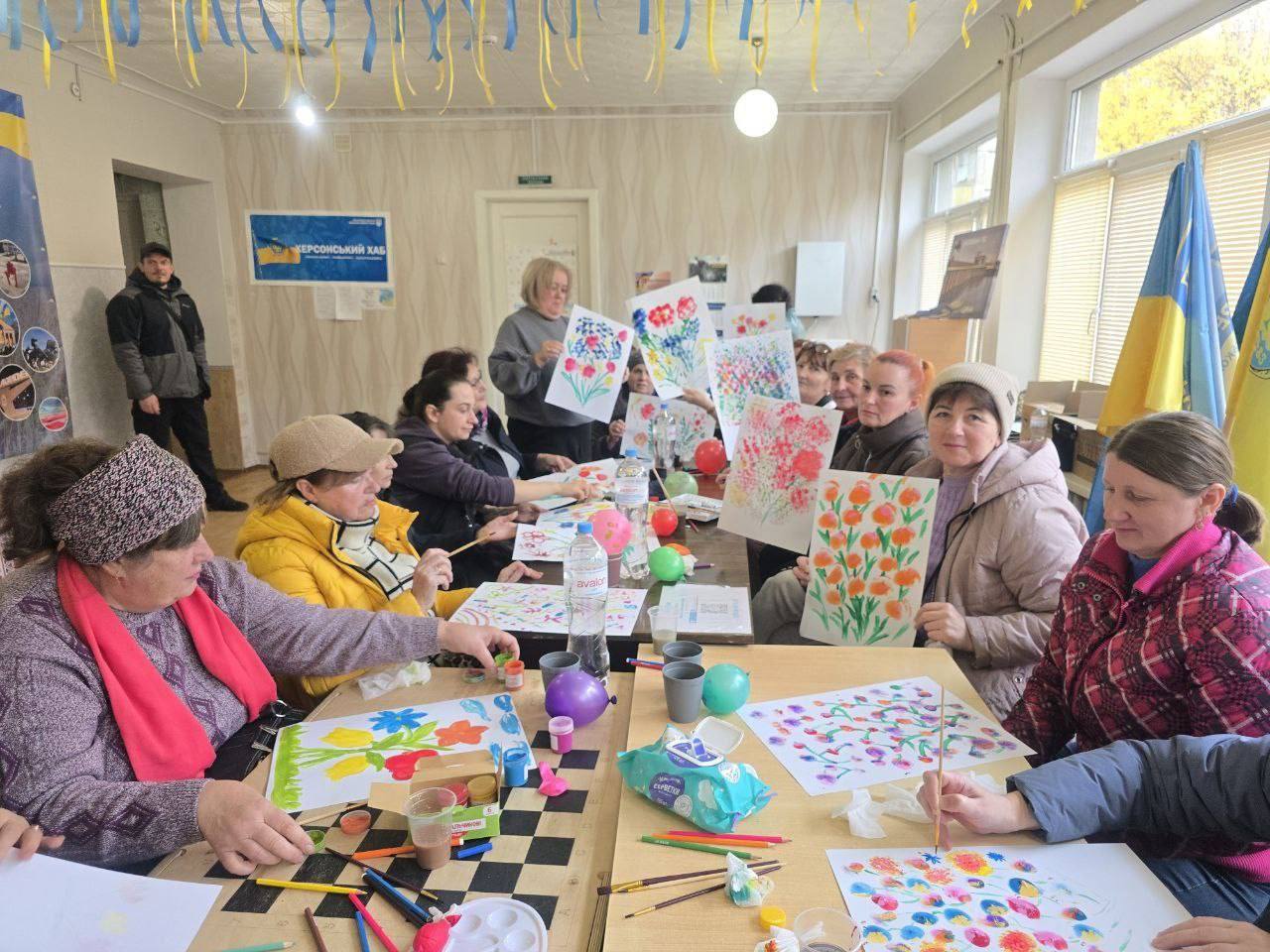 Група жінок бере участь у творчому занятті (малюванні) в приміщенні. Деякі з них тримають свої малюнки з квітами. Кімната прикрашена синьо-жовтими стрічками та прапорами України, на стіні видно банер ХЕРСОНСЬКИЙ ХАБ.