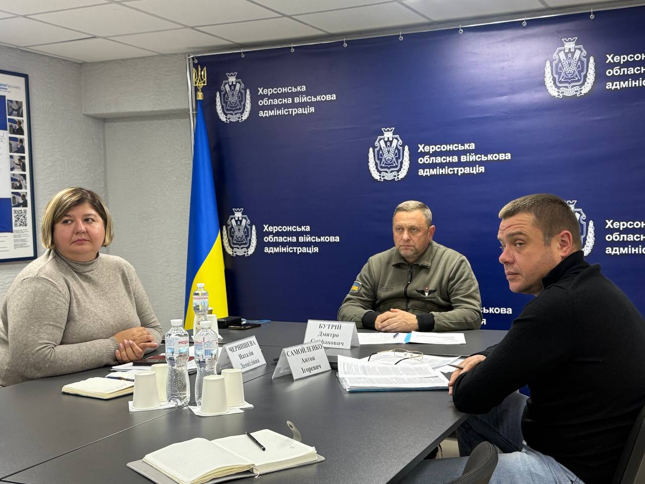 На фото троє людей (дві жінки та один чоловік у військовій формі) сидять за столом на діловій зустрічі. На задньому плані — синій банер з написом: Херсонська обласна військова адміністрація та державний прапор України.