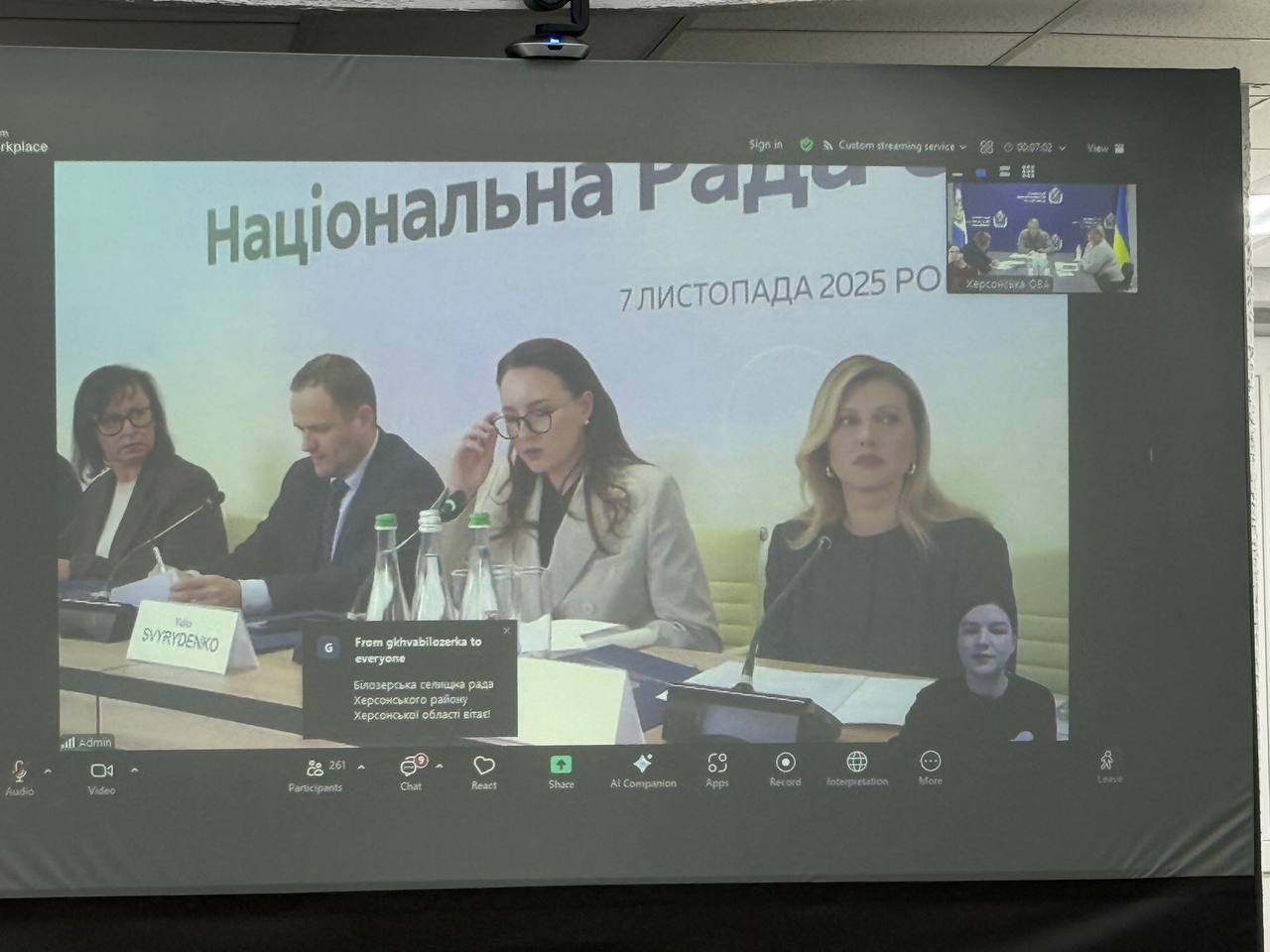 На екрані великої проекції Zoom-конференції видно чотирьох учасників, що сидять за столом та рядок субтитрів. У правому верхньому куті — менше вікно з логотипом Херсонська ОВА.