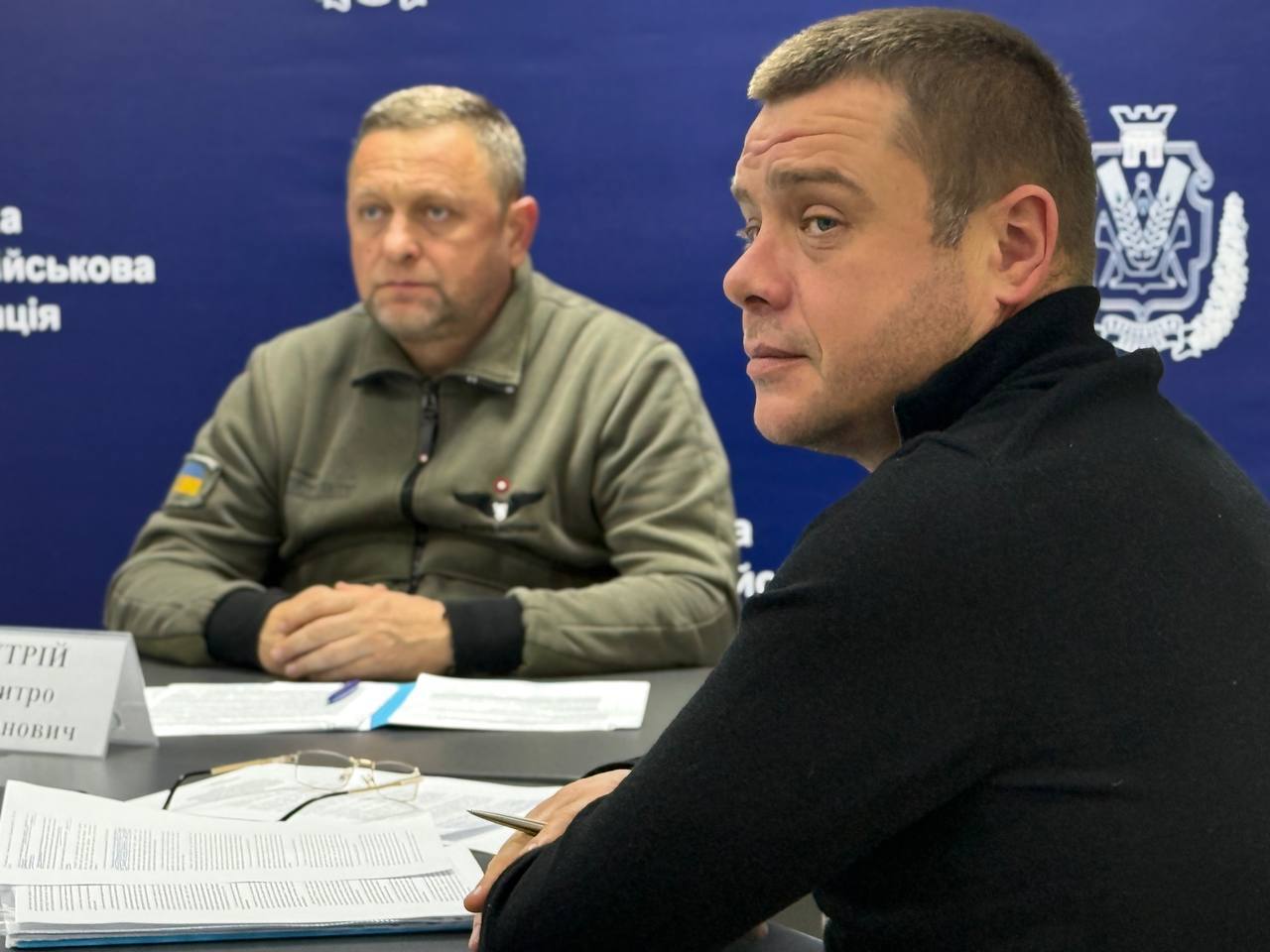 Крупний план двох чоловіків, що сидять за столом під час зустрічі. Чоловік на передньому плані, одягнений у чорний светр, дивиться праворуч, а чоловік на задньому плані, одягнений у військову форму, дивиться вперед. За ними видно синій банер.