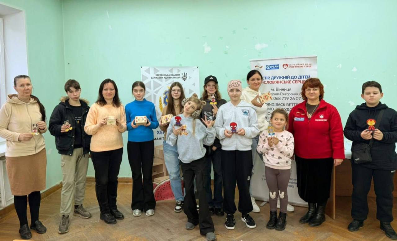 Групове фото десяти дітей та двох дорослих, які стоять у світлій кімнаті. Більшість із них тримають у руках маленькі вироби (імовірно, фігурки). На задньому плані видно банер з емблемою Херсонської обласної державної адміністрації та інформацією про благодійний фонд Слов'янське Серце.
