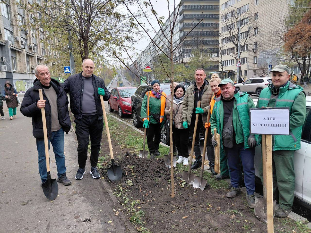 Група з дев'яти людей, переважно у робочому одязі, стоїть на міській вулиці з лопатами навколо щойно посадженого дерева. Один із учасників тримає табличку АЛЕЯ ХЕРСОНЩИНИ.