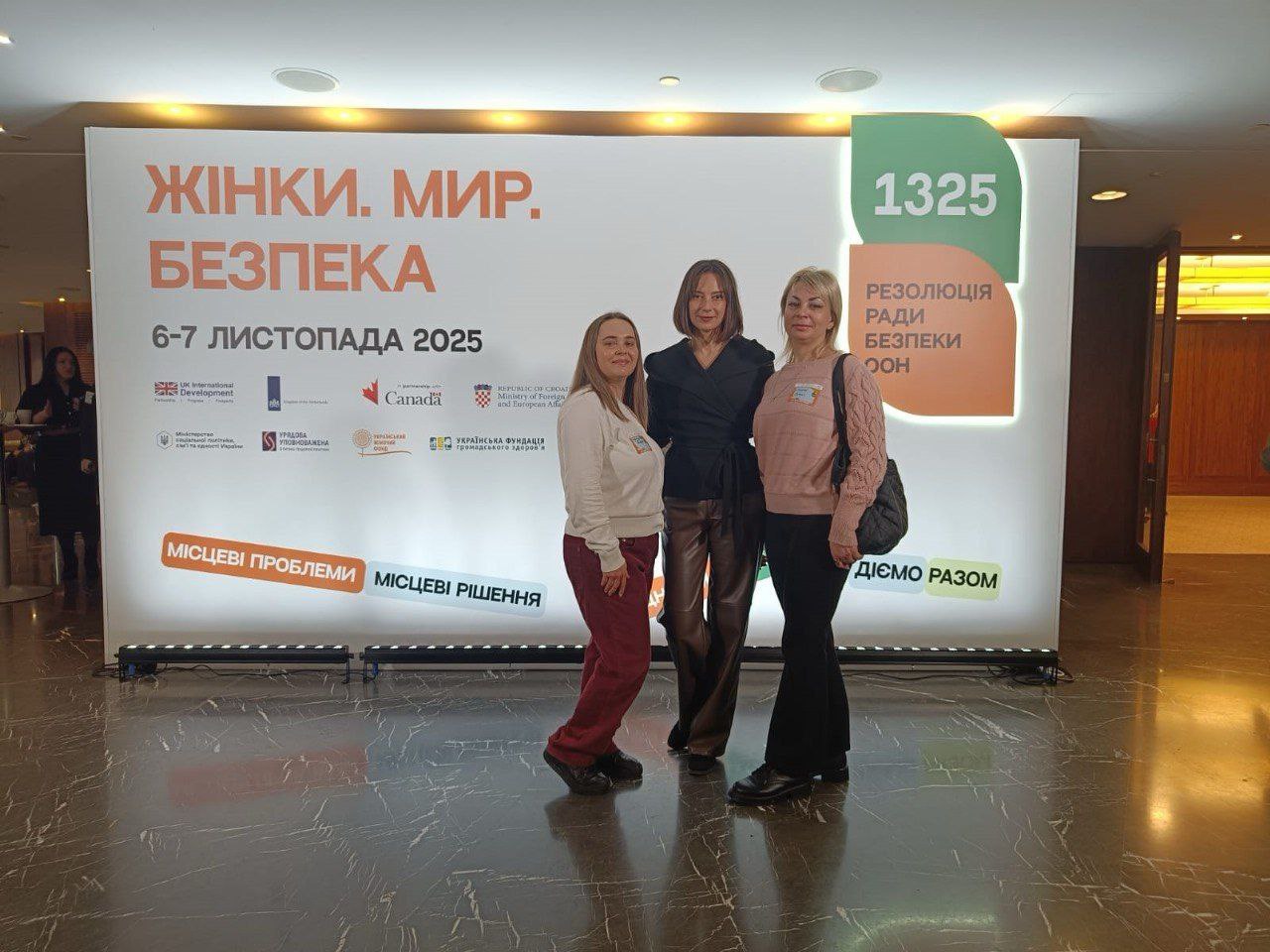 Три жінки позують перед великим банером на конференції. На банері великими літерами написано: ЖІНКИ. МИР. БЕЗПЕКА, а також: 1325 Резолюція Ради Безпеки ООН, дати 6-7 ЛИСТОПАДА 2025 та фрази: МІСЦЕВІ ПРОБЛЕМИ МІСЦЕВІ РІШЕННЯ і ДІЄМО РАЗОМ.