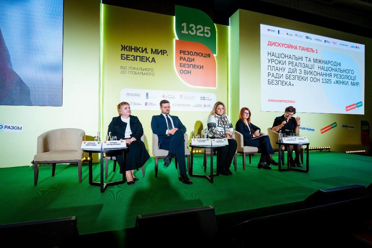 П'ятеро учасників (дві жінки ліворуч, чоловік у центрі, дві жінки праворуч) сидять на сцені під час Дискусійної панелі 1. На екранах позаду видно теми: Жінки. Мир. Безпека, 1325 Резолюція Ради Безпеки ООН та назва панелі: НАЦІОНАЛЬНІ ТА МІЖНАРОДНІ УРОКИ РЕАЛІЗАЦІЇ НАЦІОНАЛЬНОГО ПЛАНУ ДІЙ З ВИКОНАННЯ РЕЗОЛЮЦІЇ РАДИ БЕЗПЕКИ ООН 1325 ЖІНКИ. МИР. БЕЗПЕКА.