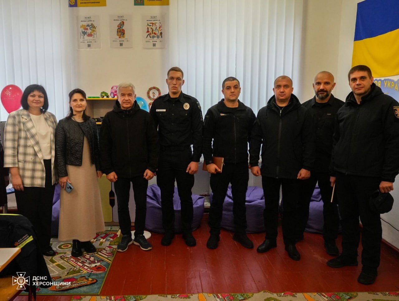 Група з семи чоловіків та двох жінок у кімнаті з навчальними плакатами та українським прапором. Чоловіки одягнені переважно у чорну форму або темний одяг, жінки – у світліший.