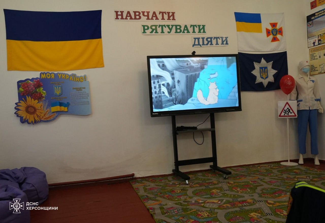 Навчальний куточок з безпеки життєдіяльності. На стіні написи: НАВЧАТИ, РЯТУВАТИ, ДІЯТИ, український прапор, прапор ДСНС України та прапор Національної поліції. У центрі інтерактивний дисплей, на якому показують мультфільм. Праворуч — манекен у медичному одязі.