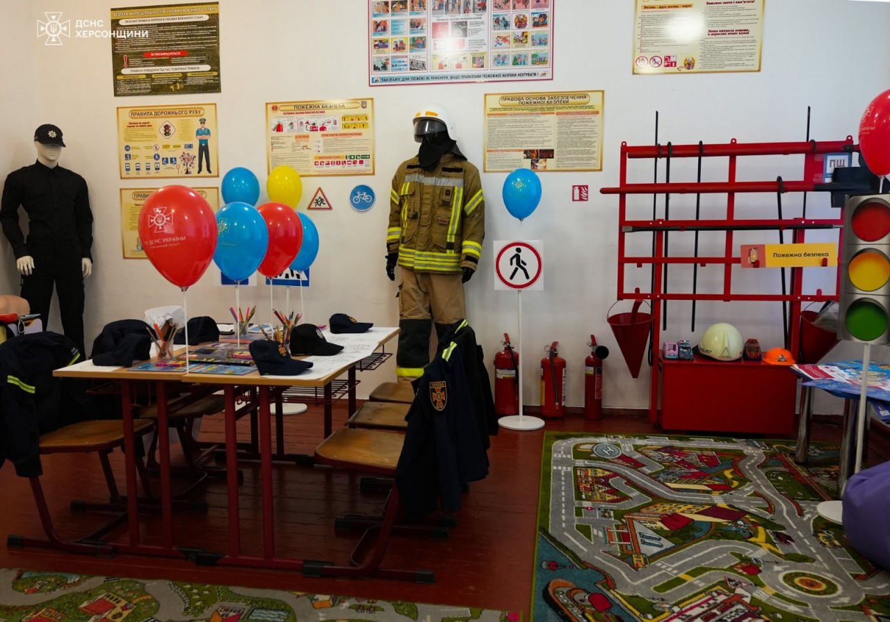Навчальний клас з безпеки, обладнаний плакатами, манекенами у формі поліцейського та пожежника, столом для занять з кепками та приладдям, а також червоними, синіми та жовтими повітряними кульками. Праворуч розташований куточок пожежної безпеки з інвентарем і великий макет світлофора.