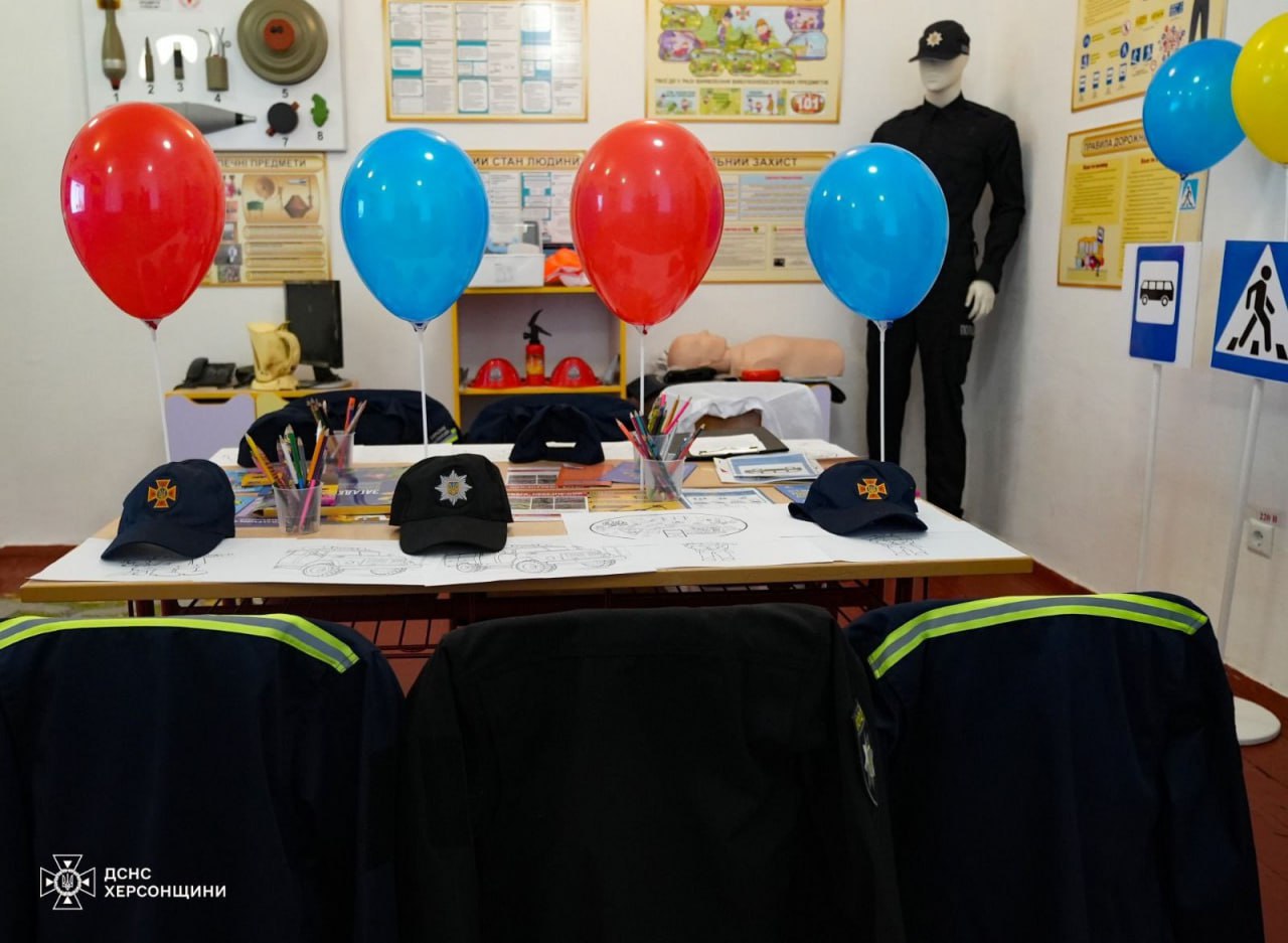 Навчальний куточок з охорони праці та безпеки життєдіяльності, прикрашений червоними та синіми повітряними кульками, трьома кепками ДСНС, розкладеним на столі приладдям для малювання та двома манекенами, один з яких одягнений у форму рятувальника.