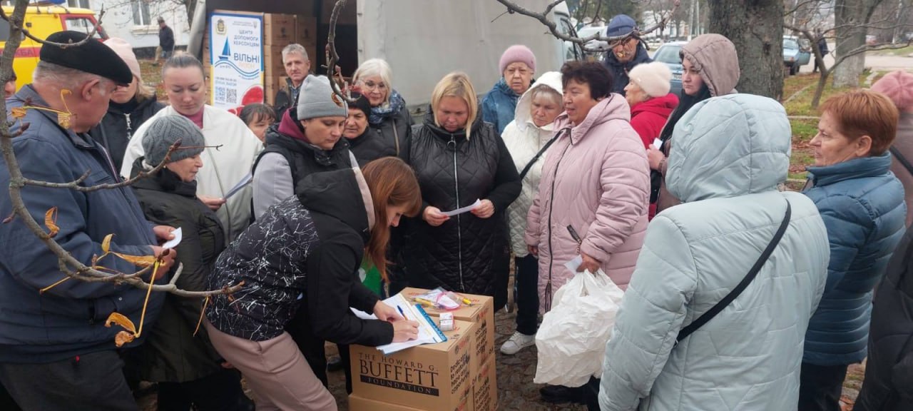 Група мешканців Херсона зібралася на вулиці для отримання гуманітарної допомоги; жінка-волонтер підписує документи на коробці з логотипом: The Howard G. Buffett Foundation на тлі стенда Центр допомоги херсонцям.