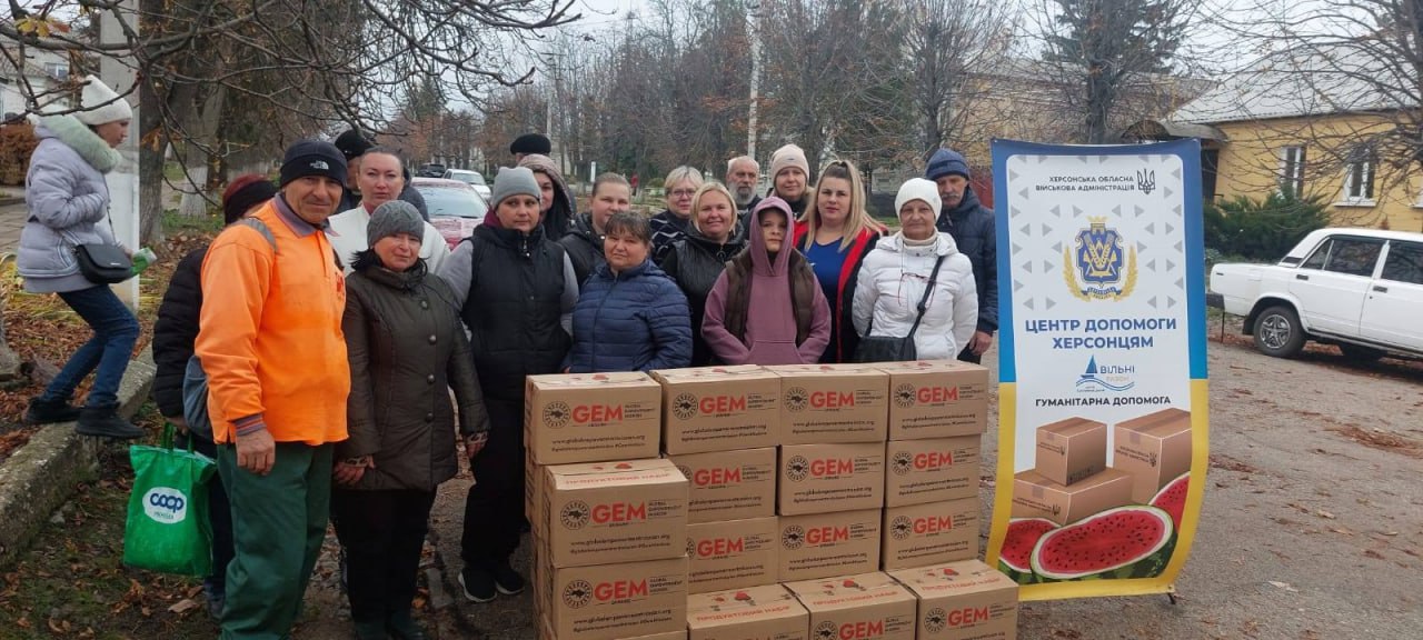 Група мешканців Херсона стоїть біля коробок із гуманітарною допомогою GEM, отриманою через Центр допомоги херсонцям (Херсонська ОВА), на тлі вулиці та житлових будинків.