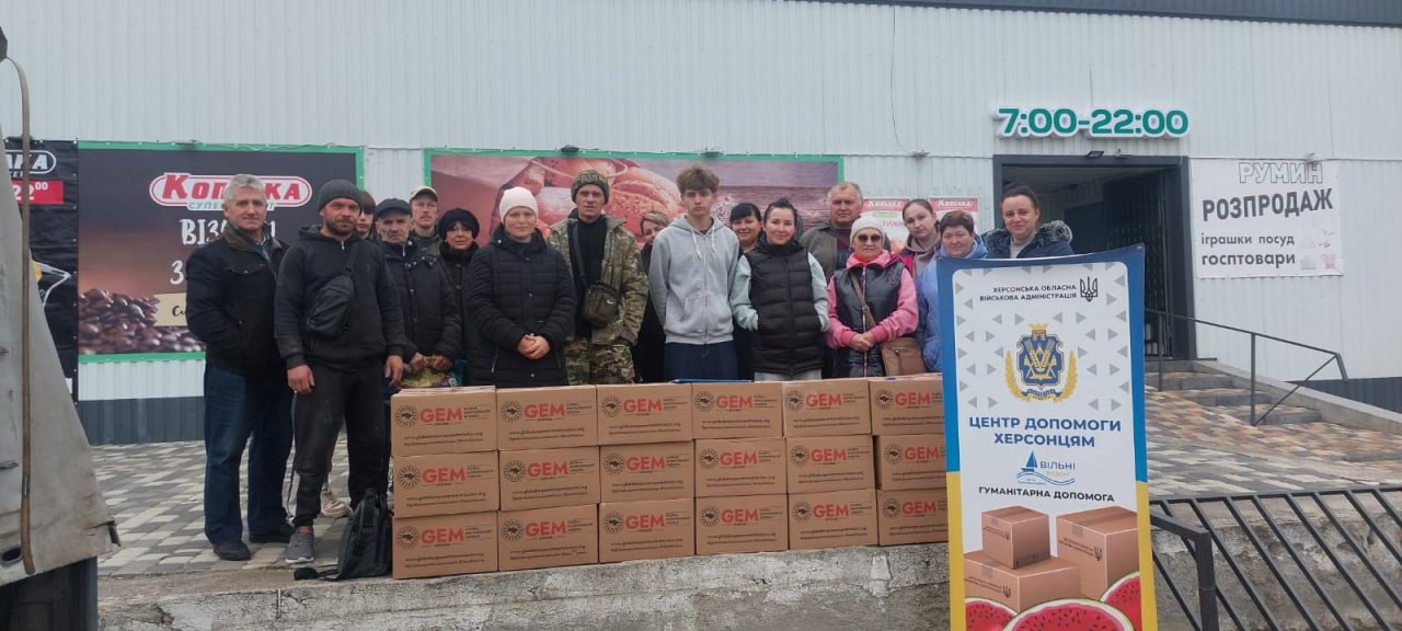 Група з близько 15 волонтерів та отримувачів стоїть перед великою кількістю коробок з гуманітарною допомогою GEM, на тлі магазину та банера Центр допомоги херсонцям.