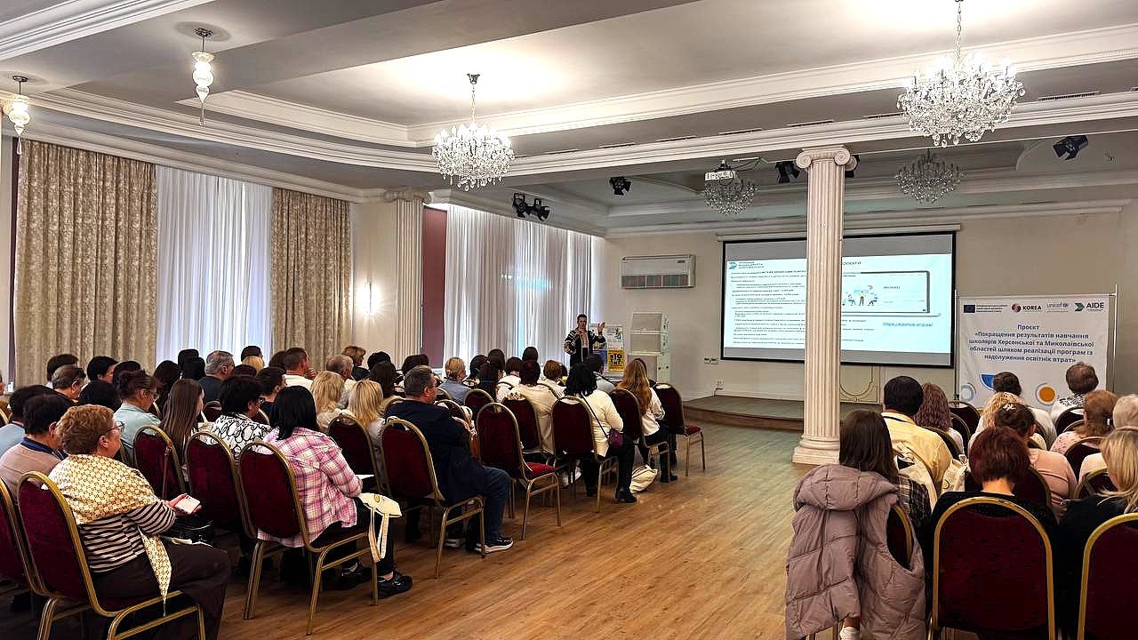 Презентація або семінар у великій залі: жінка-доповідач стоїть перед великою аудиторією, що сидить на стільцях, на тлі проекційного екрана з інформацією про проєкт, пов'язаний з Херсонською областю та освітою.