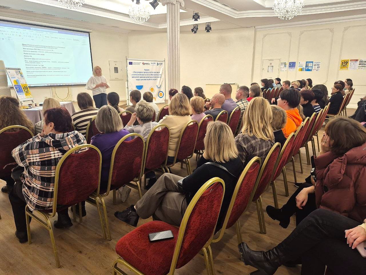 Група людей сидить у залі, слухаючи жінку-доповідача під час освітнього заходу.