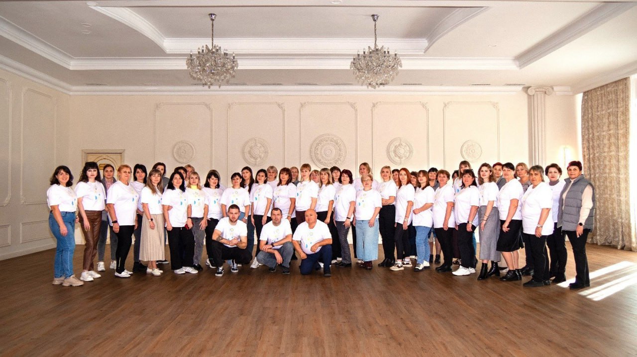 Велика група учасників (близько 50 осіб), більшість з яких одягнені в білі футболки, позують для групового фото в елегантному, просторому залі з люстрами та дерев'яною підлогою.