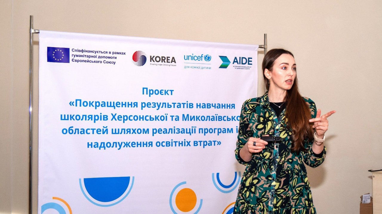 Жінка-доповідач виступає на презентації проєкту з надолуження освітніх втрат для школярів Херсонської та Миколаївської областей. На банері видно логотипи Європейського Союзу, KOREA, UNICEF та AIDE.