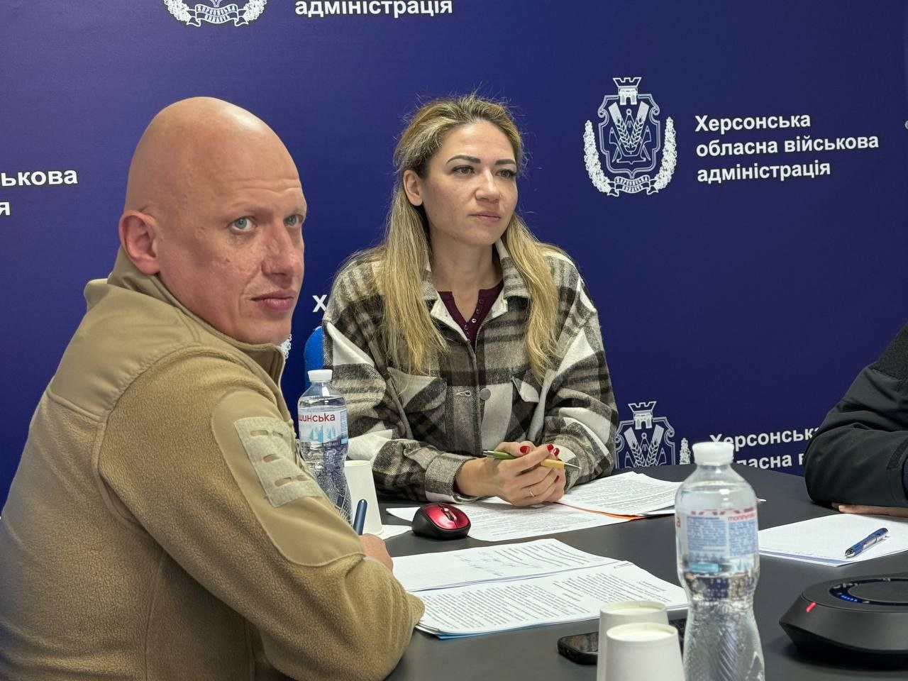 Наближений кадр двох учасників зустрічі (чоловіка у тактичній формі та жінки) за столом. На задньому плані — логотип Херсонської обласної військової адміністрації.