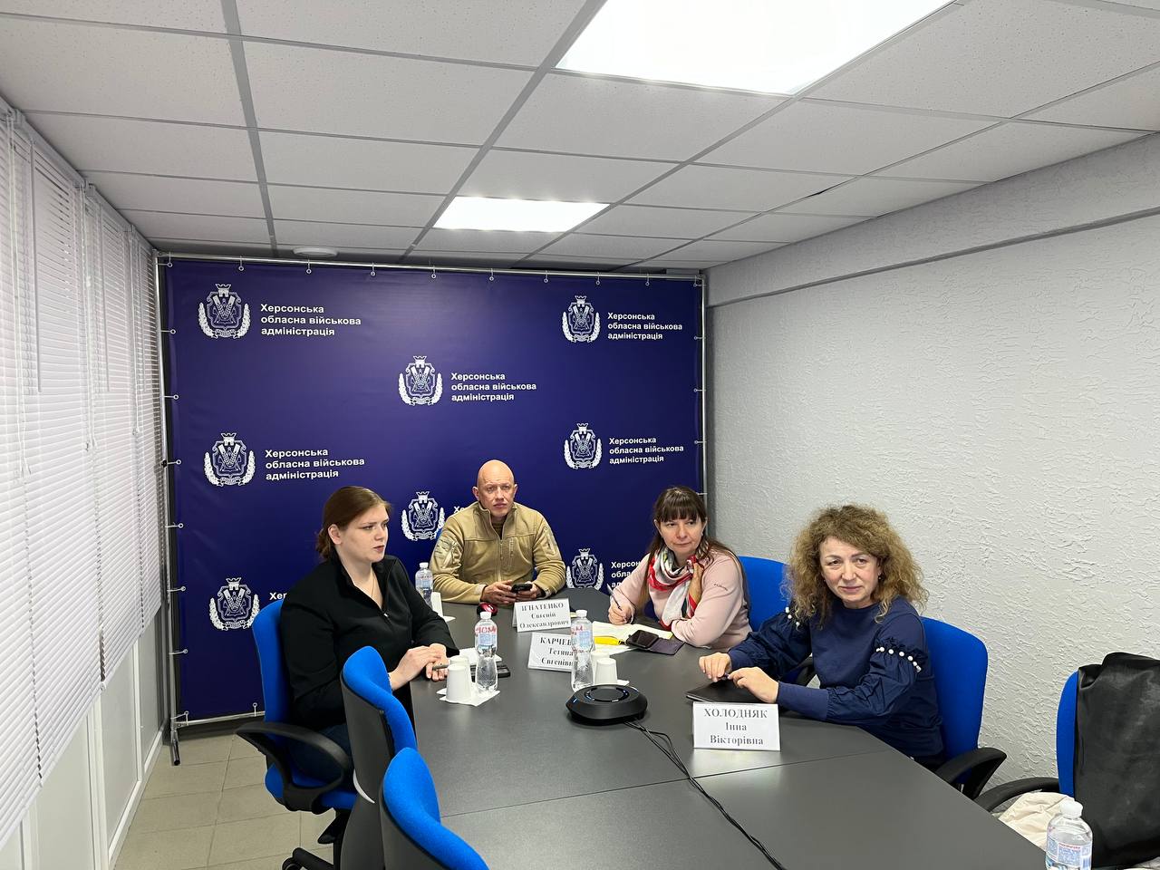 Групове фото чотирьох людей (двох жінок і двох чоловіків) за столом у приміщенні на фоні стіни з логотипами Херсонської обласної військової адміністрації. На столі видно пляшки з водою та мікрофон.