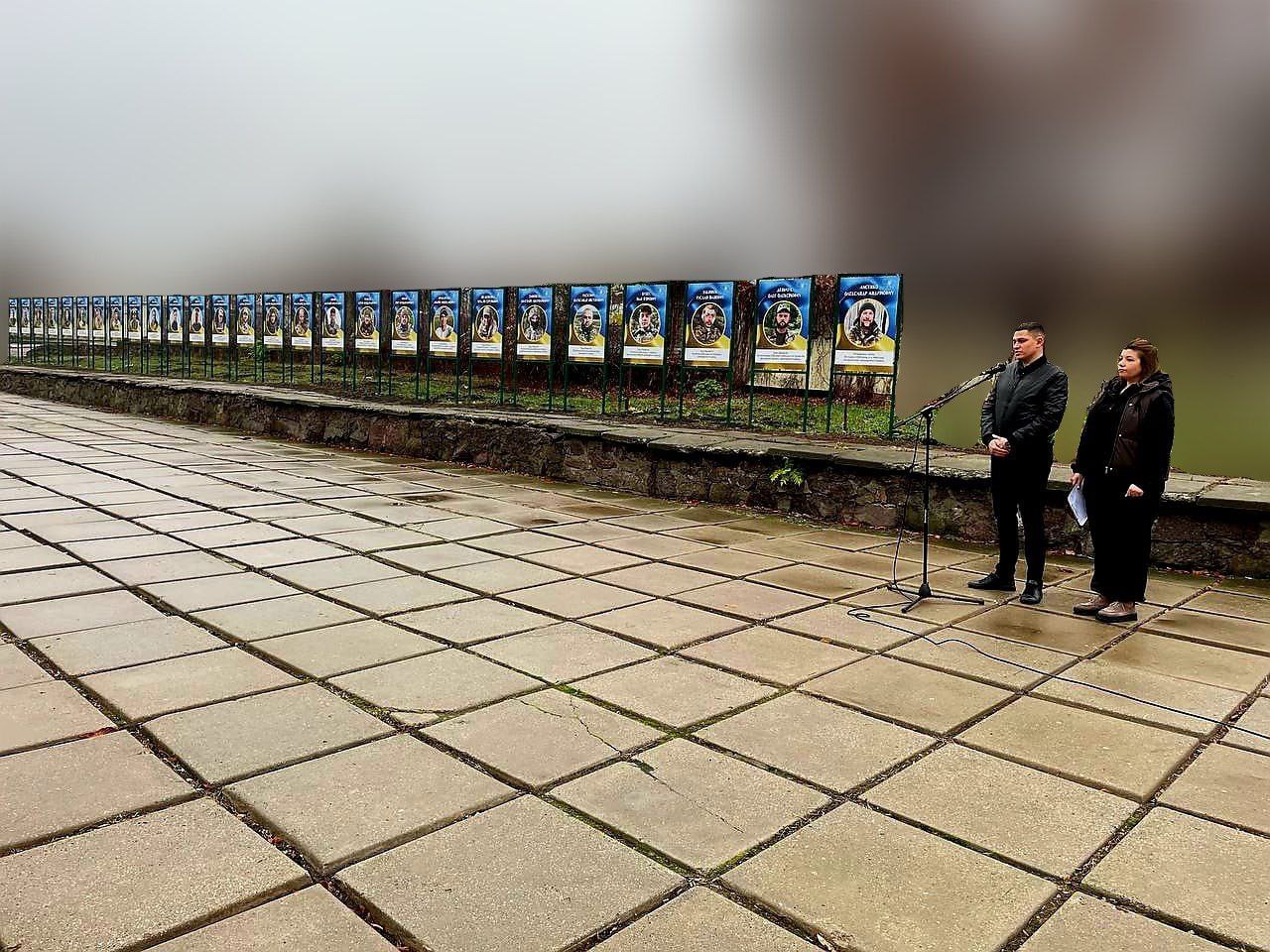Меморіальна виставка портретів на алеї, де двоє людей виступають перед мікрофоном на тлі низки меморіальних стендів під час похмурої погоди.