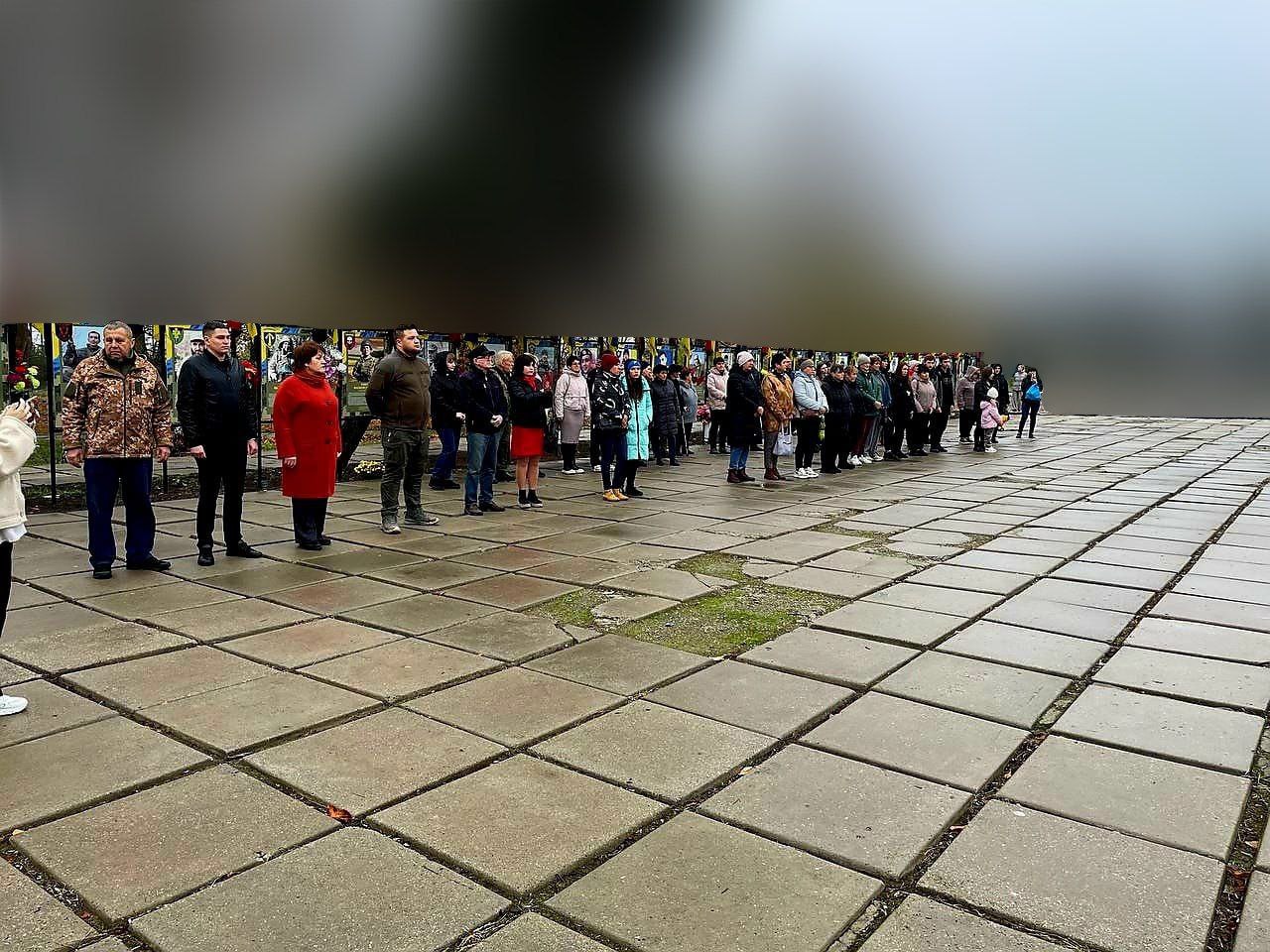 Група людей стоїть на площі з плит, беручи участь у церемонії біля стендів із портретами українських захисників.