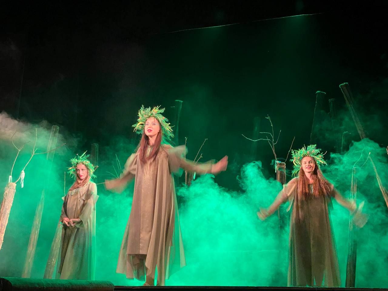 Три актриси в довгих зеленувато-коричневих сукнях та вінках із колосків або листя стоять на сцені. Сцена освітлена зеленим світлом, навколо них клубочиться дим, створюючи містичну атмосферу, а на задньому плані видно декорації з гілок і стовбурів.
