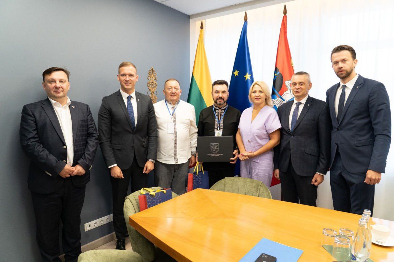 Групова фотографія, на якій сім осіб (шість чоловіків і одна жінка) позують у приміщенні біля столу для переговорів. На задньому плані видно прапори: Литви, Європейського Союзу та України, а також прапор з гербом.