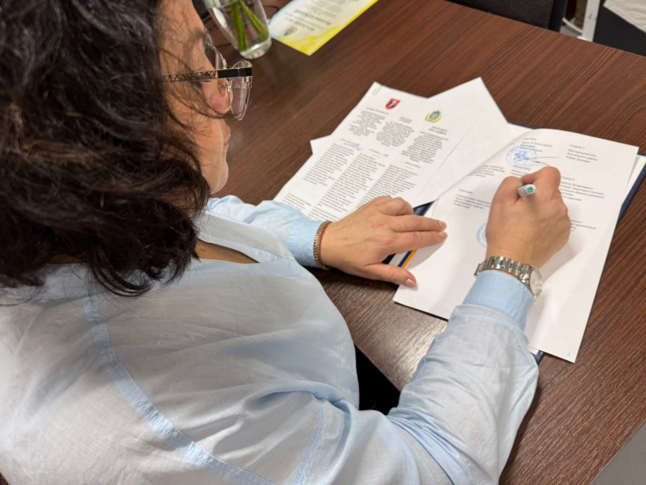 Жінка в окулярах і світло-блакитній сорочці сидить за столом і підписує ручкою документ, який лежить на столі разом з іншими паперами. Знімок зроблено збоку, зверху, фокусуючись на її руках і документах.