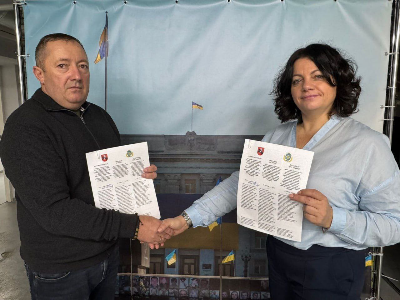 Двоє людей – чоловік і жінка – стоять і потискають один одному руки, тримаючи вільними руками документи. Вони знаходяться перед фоном із зображенням будівлі, українського прапора та фотографій загиблих захисників.