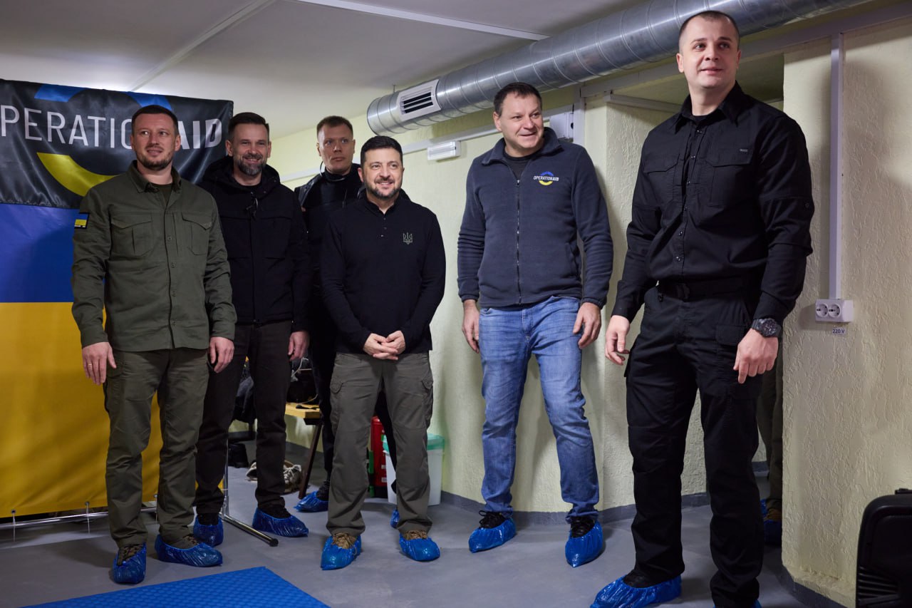 Групове фото Президента України Володимира Зеленського та його супроводу, які стоять у приміщенні на синіх бахілах. На задньому плані видно частину банера з написом OPERATION AID та український прапор.