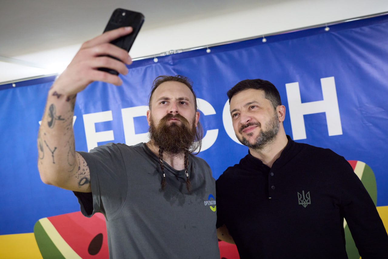Селфі Президента України Володимира Зеленського (в чорній кофті) з чоловіком з довгою бородою та татуюваннями, який тримає телефон. На задньому плані видно синій банер з написом: ХЕРСОН та зображенням кавуна.
