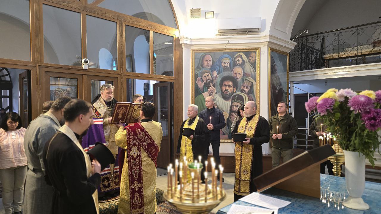Релігійна служба проходить у приміщенні церкви, де кілька священників у літургійному вбранні тримають книги та свічки. Поруч стоять парафіяни та військовослужбовець. На задньому плані видно велику ікону, а на передньому плані — вазу з квітами та свічник із запаленими свічками.