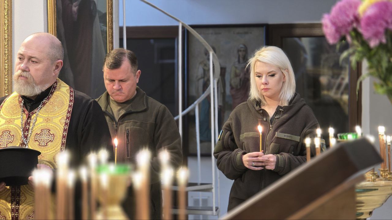 Православний священник у літургійному вбранні стоїть поруч із двома парафіянами – чоловіком у зеленій формі та білявою жінкою також у формі, яка тримає запалену свічку. Усі троє зосереджені, беручи участь у службі, на передньому плані видно запалені свічки у свічнику, а на задньому — ікони та гвинтові сходи.
