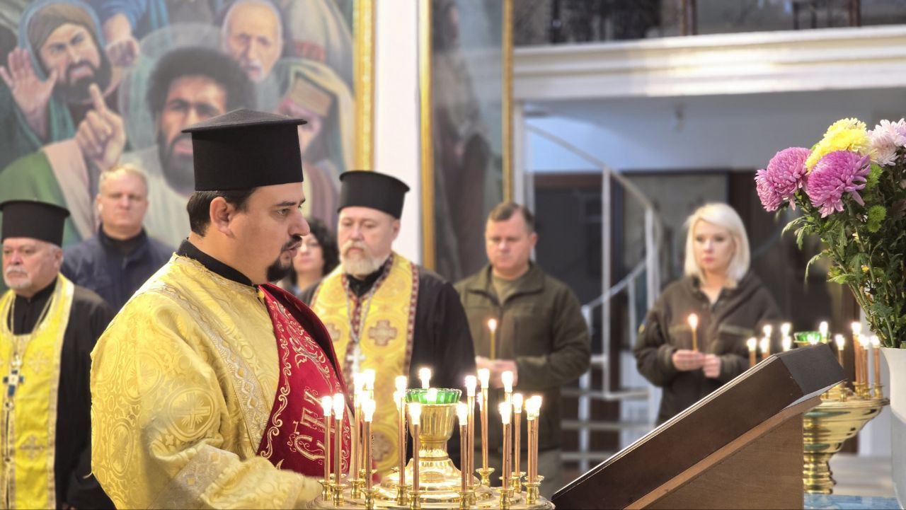 Священник у золотистому літургійному вбранні та чорній камилавці стоїть перед свічником, очолюючи релігійну службу. Навколо нього стоять інші священники та парафіяни, деякі з яких тримають свічки. На задньому плані видно частину великої ікони та букет квітів справа.