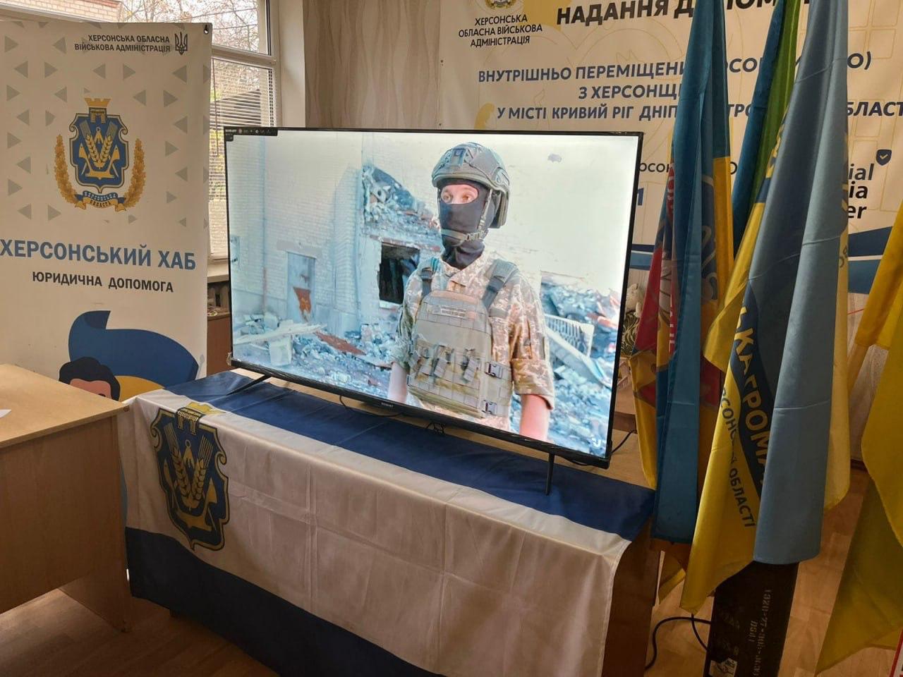 Телевізор у центрі Херсонський Хаб. Юридична допомога показує зображення українського військового у масці та бронежилеті на тлі зруйнованих будівель. Сцена обрамлена українськими прапорами та банерами Херсонської ОВА.