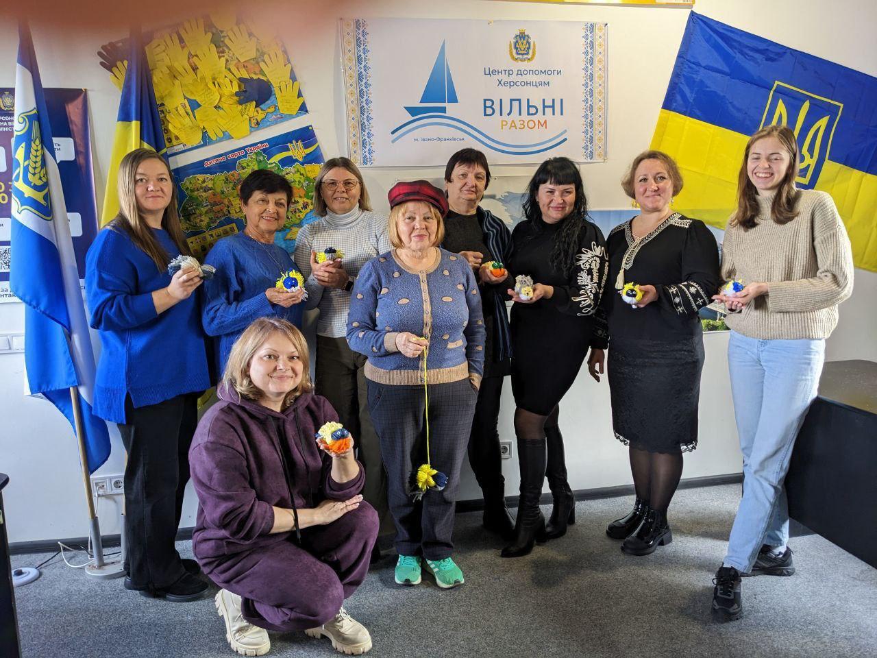 Група з дев'яти жінок посміхається, тримаючи маленькі сувеніри, схожі на брелоки або помпони, у синьо-жовтих кольорах. Жінки стоять на тлі українських прапорів та банера Центр допомоги херсонцям. Вільні разом у місті Івано-Франківськ.