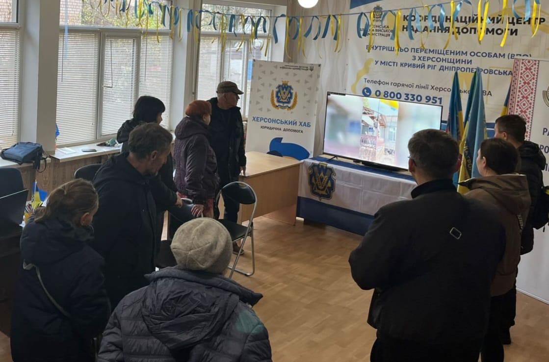 Група людей зібралася навколо телевізора в кімнаті, прикрашеній українськими прапорами та символікою Херсонський Хаб. Належна допомога. На екрані телевізора демонструється відео, а також видно номер телефону гарячої лінії.