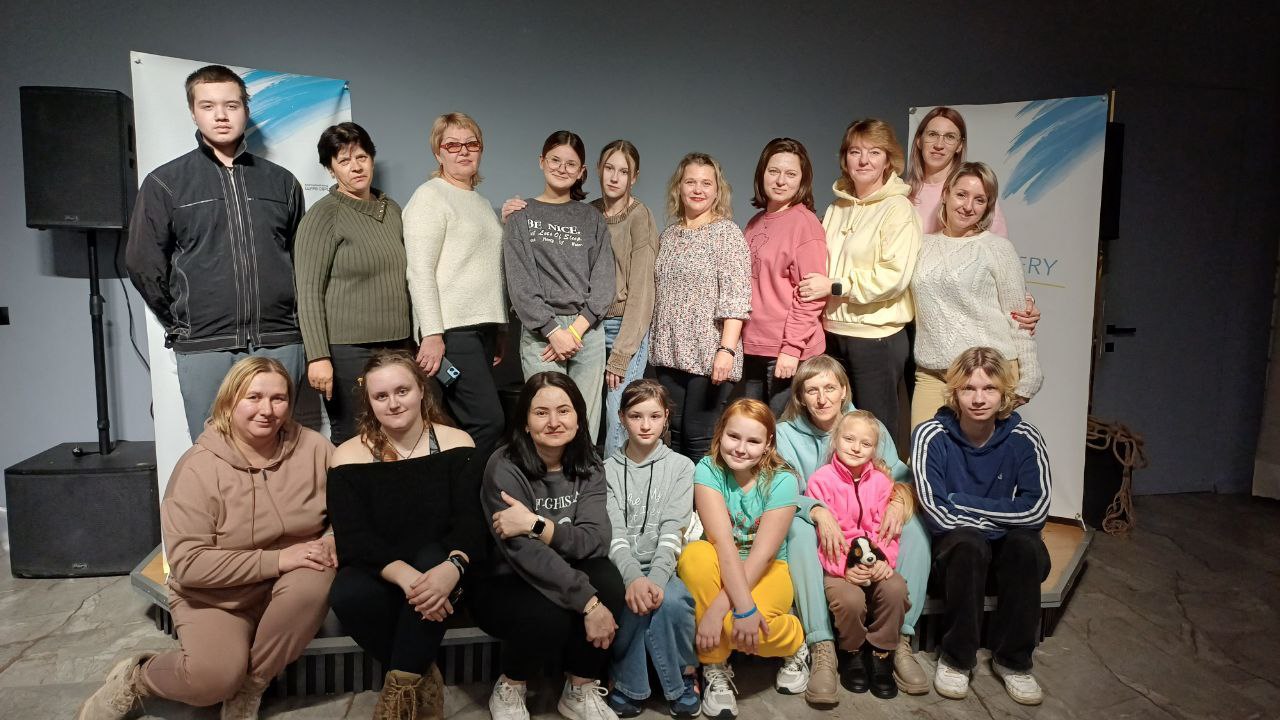 Групова фотографія, на якій зображено приблизно 18 людей – переважно жінки та дівчата, а також двоє хлопців – позують у приміщенні. Люди стоять або сидять у два ряди, на тлі видно дві великі вертикальні панелі з блакитними акцентами.