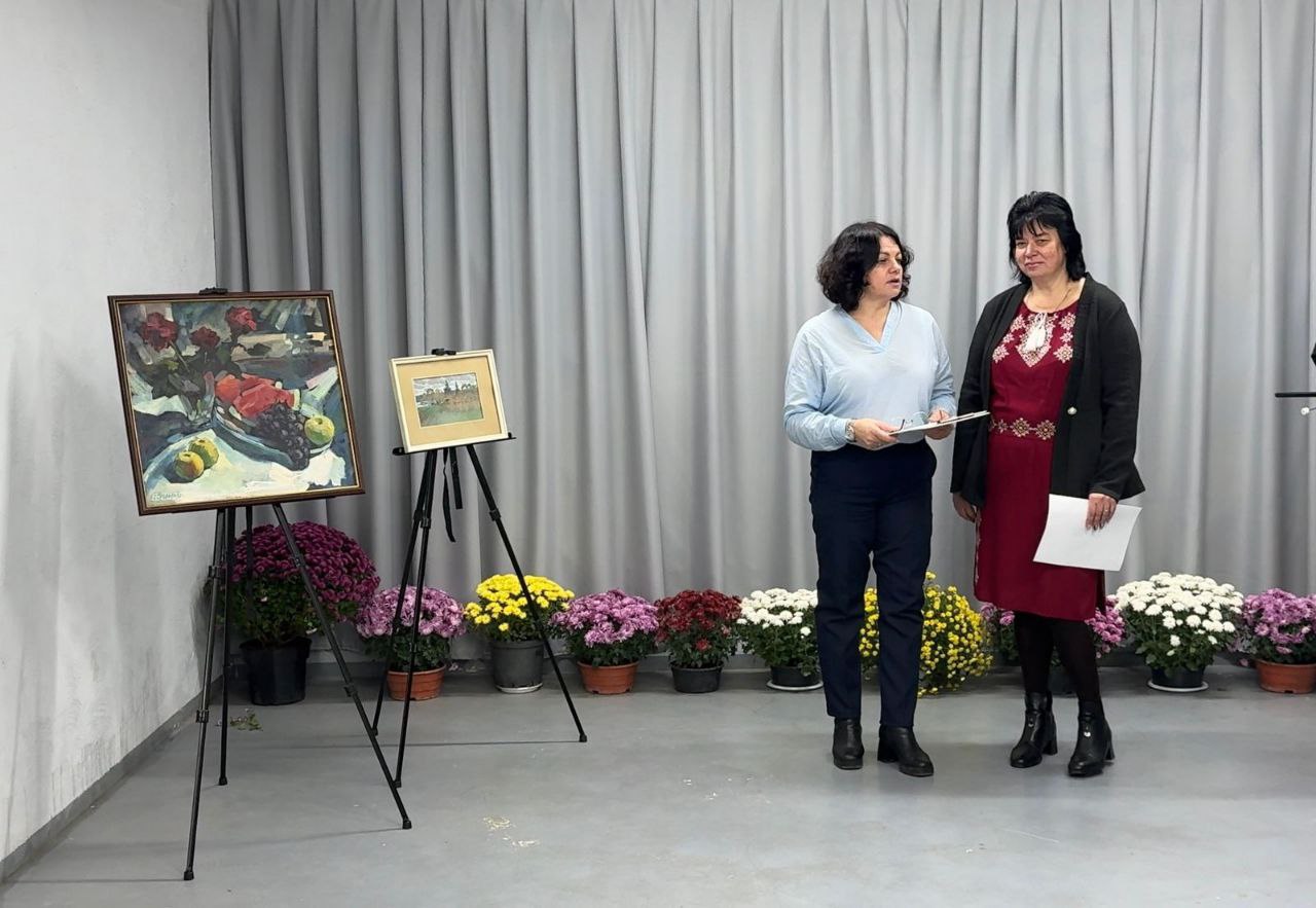 Дві жінки стоять у приміщенні перед сірою драпіровкою під час виставки чи презентації. Зліва розташовані мольберти з картинами, а вздовж стіни — горщики з різнокольоровими хризантемами (жовтими, білими, бордовими та фіолетовими).