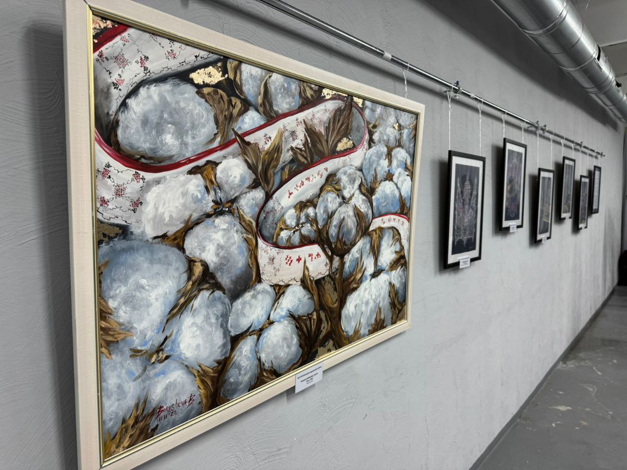 Фрагмент художньої виставки в приміщенні: на світло-сірій стіні висить велика картина з абстрактним зображенням великих, пухнастих білих форм, схожих на бавовну, з коричневими елементами та червоно-білою стрічкою. Праворуч уздовж стіни підвішено в ряд кілька менших, темних картин у рамках.