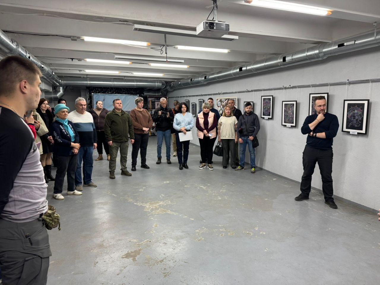 Груповий знімок з виставки або презентації в галерейному просторі. Близько п'ятнадцяти відвідувачів стоять по центру, уважно слухаючи чоловіка в чорному одязі, який стоїть праворуч і жестикулює рукою до підборіддя. На стіні позаду відвідувачів висить ряд невеликих картин у темних рамках.