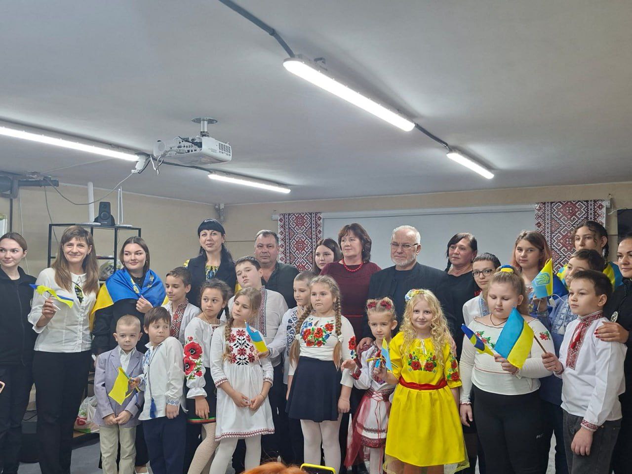 Велика група дорослих та дітей у вишиванках і з невеликими українськими прапорцями, стоять разом у приміщенні з низькою стелею.