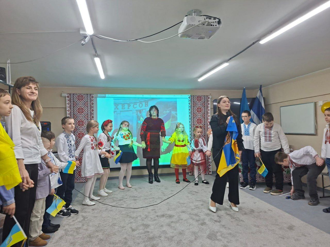 Діти у вишиванках та національному одязі, що тримаються за руки, утворюючи півколо навколо жінки в центрі; на задньому плані проекційний екран зі словом: ХЕРСОН.