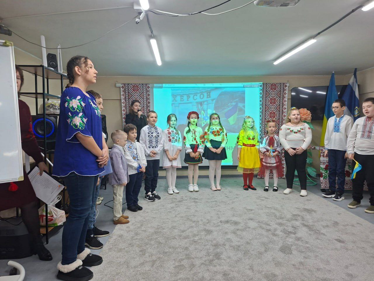 Група дітей у вишиванках стоїть півколом перед проекційним екраном зі словом: ХЕРСОН під час виступу, поруч стоїть доросла жінка.