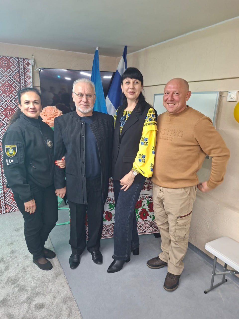 Чотири дорослі особи — двоє чоловіків і дві жінки, одна з яких у поліцейській формі, а інша у вишиванці — стоять разом для групового фото на тлі українського орнаменту та прапорів.