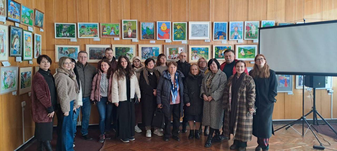 Групове фото людей, що стоять у приміщенні з дерев'яною обшивкою стін, на тлі виставки дитячих малюнків і великого проекційного екрана.